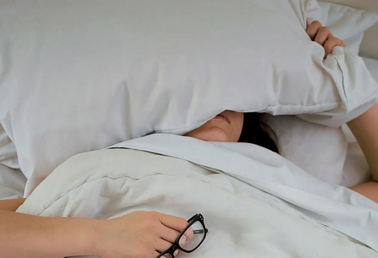 person lying on bed while covering face with pillow and holding eyeglasses